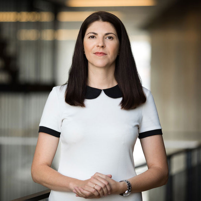 Portret kobiety w białej sukience z czarnym kołnierzykiem i mankietami, z dłońmi splecionymi na wysokości talii, w tle jasne wnętrze biurowe.