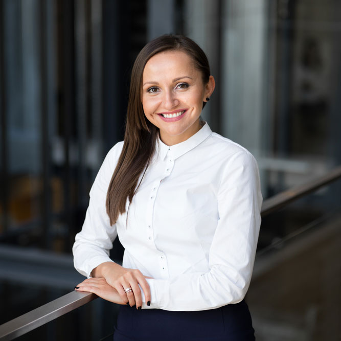 Portret uśmiechniętej kobiety w białej koszuli, opierającej się o metalową barierkę na tle nowoczesnego biurowca.