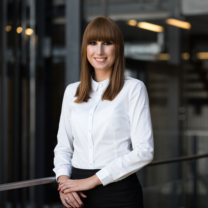 Portret uśmiechniętej adwokatki Alicji Krzyśko-Makulskiej w białej koszuli i czarnej spódnicy, opierającej się o metalową barierkę na tle nowoczesnego biura.
