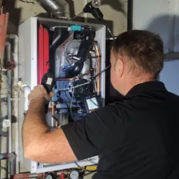 Mężczyzna w czarnej koszulce serwisuje piec gazowy, używając dwóch lamp inspekcyjnych i miernika. Widoczne rury miedziane i elementy instalacji grzewczej na ścianie.