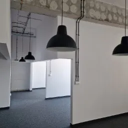 Wnętrze biurowe z trzema czarnymi lampami wiszącymi, odsłoniętym betonowym stropem z widocznymi instalacjami i białymi ścianami z czarnymi kablami elektrycznymi.