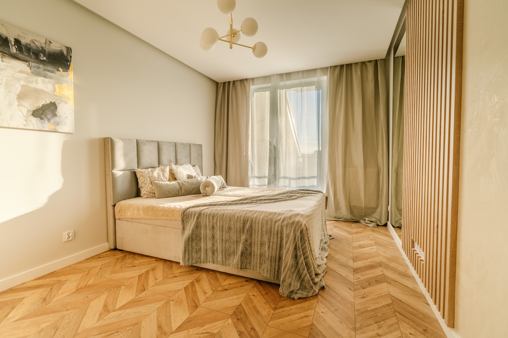 Jasna sypialnia z parkietem w jodełkę, łóżkiem z szarym zagłówkiem i beżową narzutą, obraz abstrakcyjny z elementami złota na ścianie i designerska lampa sufitowa