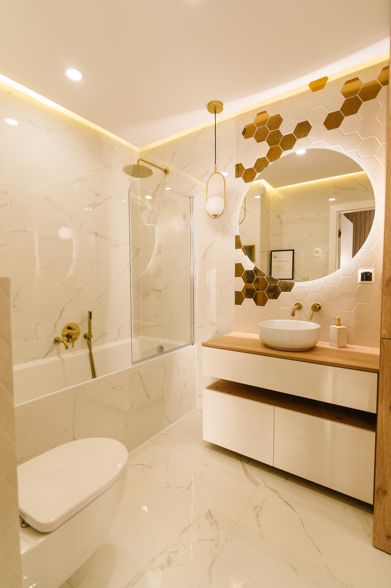 Jasna łazienka z marmurowymi płytkami, złotymi akcentami, okrągłym lustrem z podświetleniem, wiszącą lampą, białą umywalką nablatową i armaturą w kolorze złota.