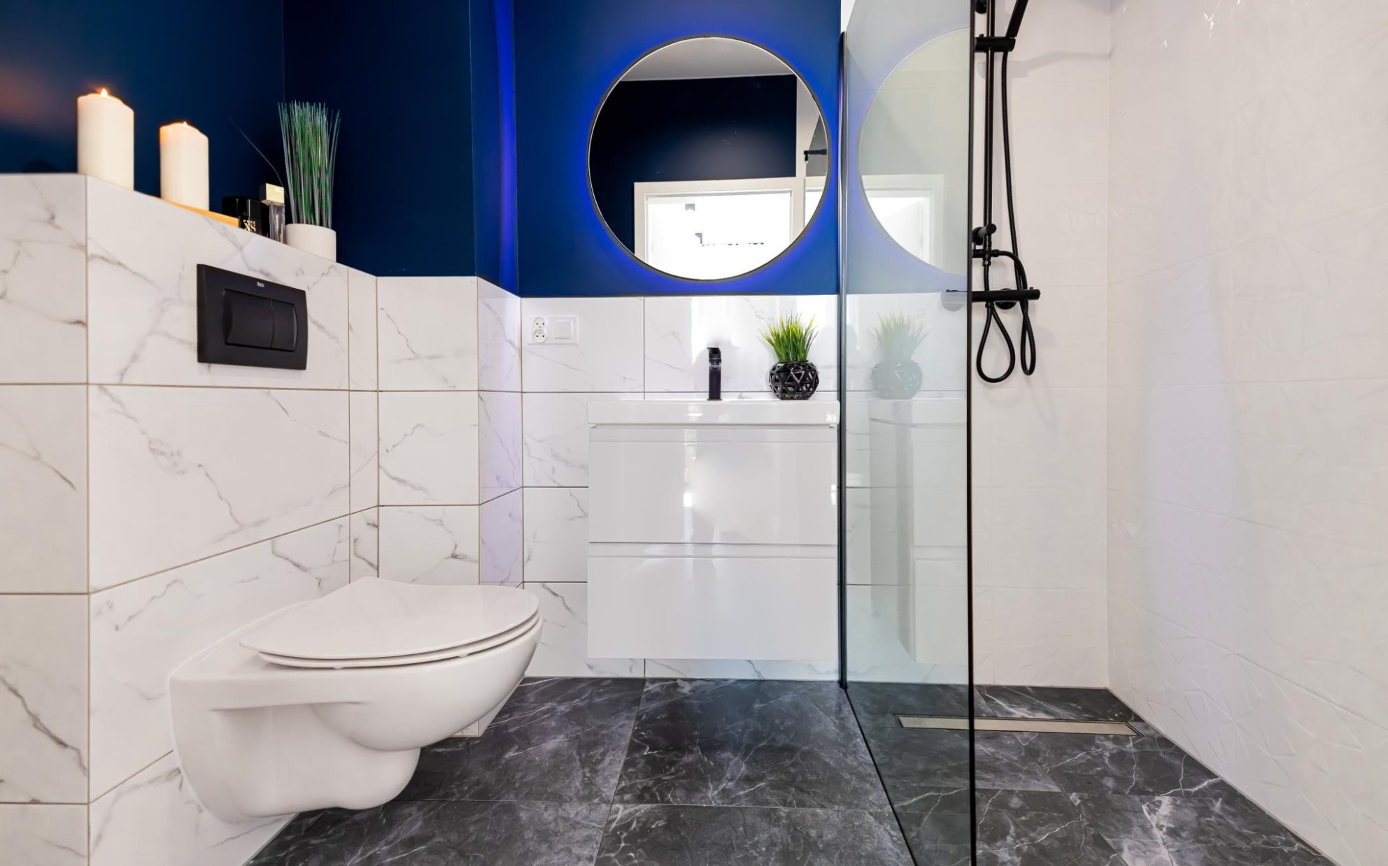 Nowoczesna łazienka z marmurowymi płytkami, podwieszaną toaletą, okrągłym lustrem z podświetleniem LED, czarną armaturą i kabiną prysznicową ze szklaną ścianką.