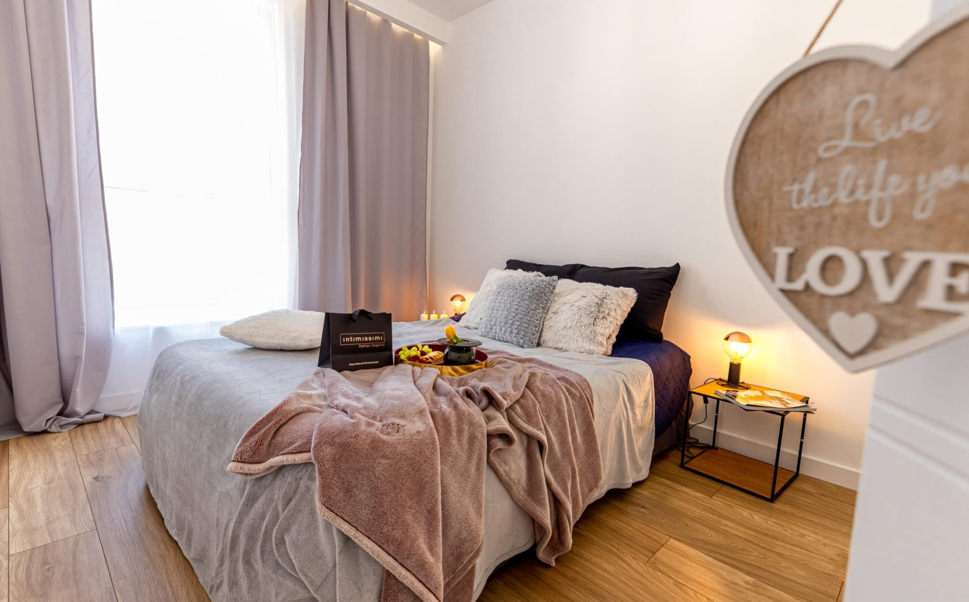 Przytulna sypialnia z łóżkiem nakrytym szarym pledem, różowym kocem i dekoracyjną tacą z owocami, oświetlona lampkami nocnymi i świecami, z drewnianym sercem z napisem 'Live the life you love'...