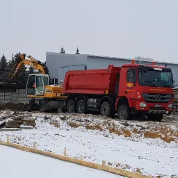 Żółta koparka Liebherr ładuje urobek do czerwonej ciężarówki na zasypanym śniegiem placu budowy z widocznymi fundamentami.