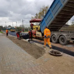 Układanie asfaltu na drodze z kostki betonowej przy użyciu rozściełacza Dynapac i ciężarówki z otwartą skrzynią ładunkową; robotnicy w odblaskowych ubraniach wyrównują nawierzchnię.
