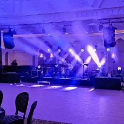 Sala balowa z przygotowaną sceną oświetloną niebieskim światłem, widoczne instrumenty muzyczne, kolumny głośnikowe, stoliki z nakryciami dla gości.