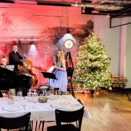 Wigilia w restauracji: wokalistka śpiewa przy mikrofonie obok pianina i kontrabasu, w tle świąteczna choinka i dekoracyjne oświetlenie punktowe, nakryty stół w pierwszym planie.