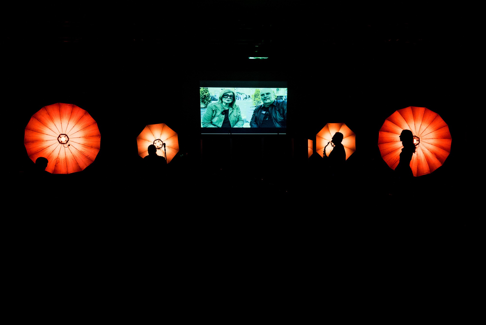 Scena z ekranem wyświetlającym parę, otoczona dużymi, okrągłymi lampami w kształcie parasoli, podświetlającymi sylwetki osób.