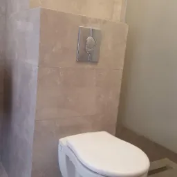 Podwieszana toaleta z chromowanym przyciskiem spłukującym, zamontowana na ścianie wyłożonej beżowymi płytkami imitującymi marmur w łazience w Poznaniu. Widoczny odpływ liniowy w podłodze.