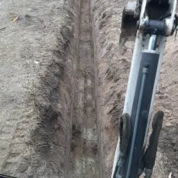 Wąski wykop w ziemi, widok z kabiny koparki, zabezpieczony taśmą ostrzegawczą, obok droga asfaltowa.