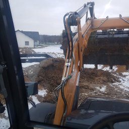 SP.mr - Widok z kabiny koparko-ładowarki CASE 695 podczas pracy w terenie z resztkami śniegu, w tle widoczny dom jednorodzinny, łyżka koparki pełna ziemi.