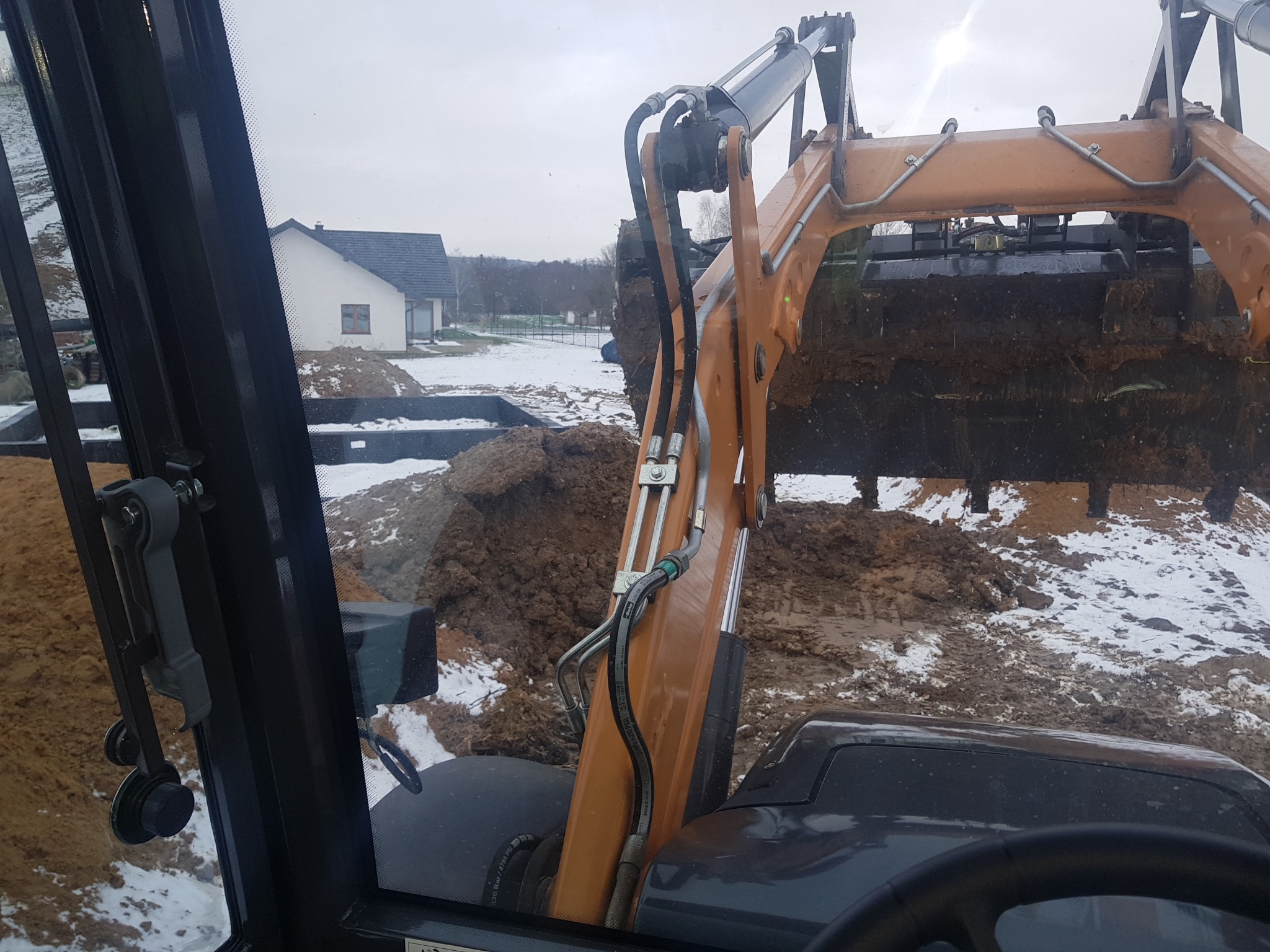 Widok z kabiny koparko-ładowarki CASE 695 podczas pracy w terenie z resztkami śniegu, w tle widoczny dom jednorodzinny, łyżka koparki pełna ziemi.