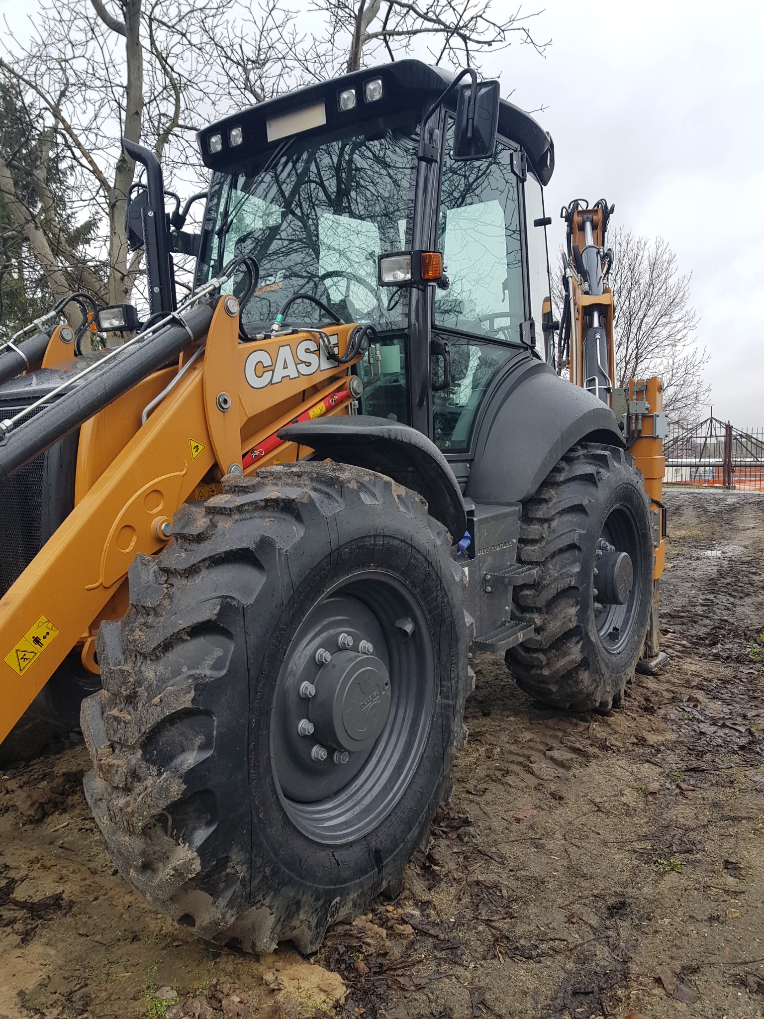Żółto-szara koparko-ładowarka marki CASE stoi na błotnistym terenie, z widocznymi oponami pokrytymi błotem i elementami konstrukcyjnymi. W tle widoczne drzewa bez liści i fragment ogrodzenia.