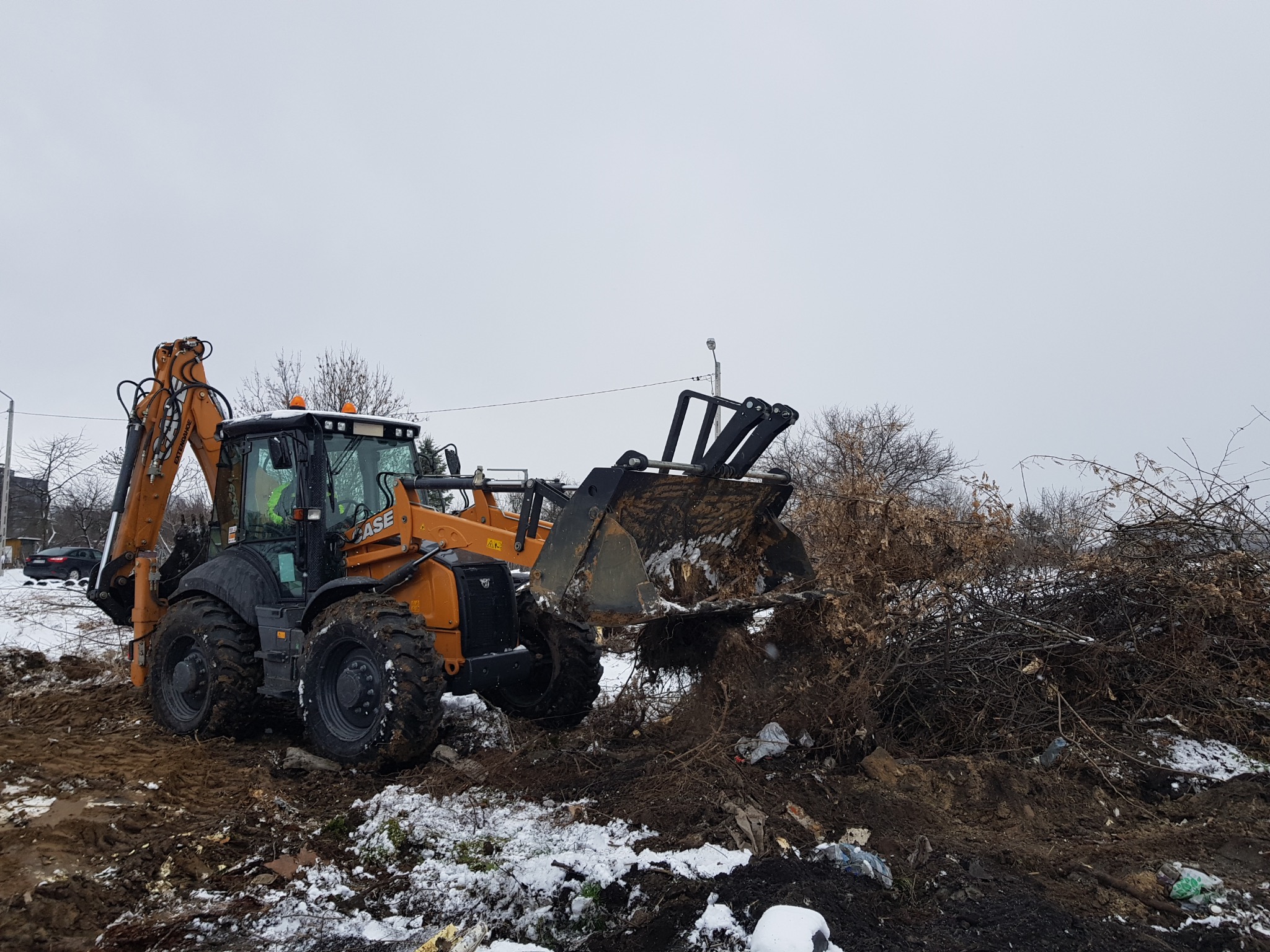 Żółto-czarna koparko-ładowarka CASE podczas prac ziemnych, przenosząca gałęzie i resztki roślinności na zasypanym śniegiem terenie.