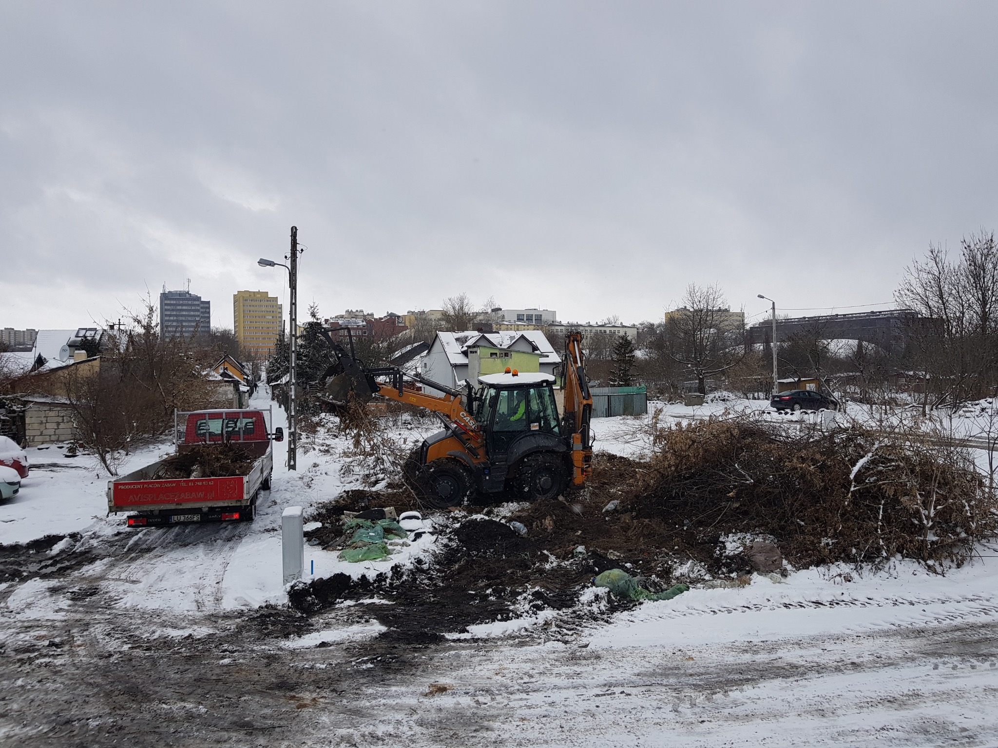 Pomarańczowo-czarna koparko-ładowarka Case usuwa gałęzie i pozostałości po wycince drzew, które są ładowane na czerwoną ciężarówkę z napisem 'PRODUCENT PLACÓW ZABAW' w zimowej scenerii z budynkami...