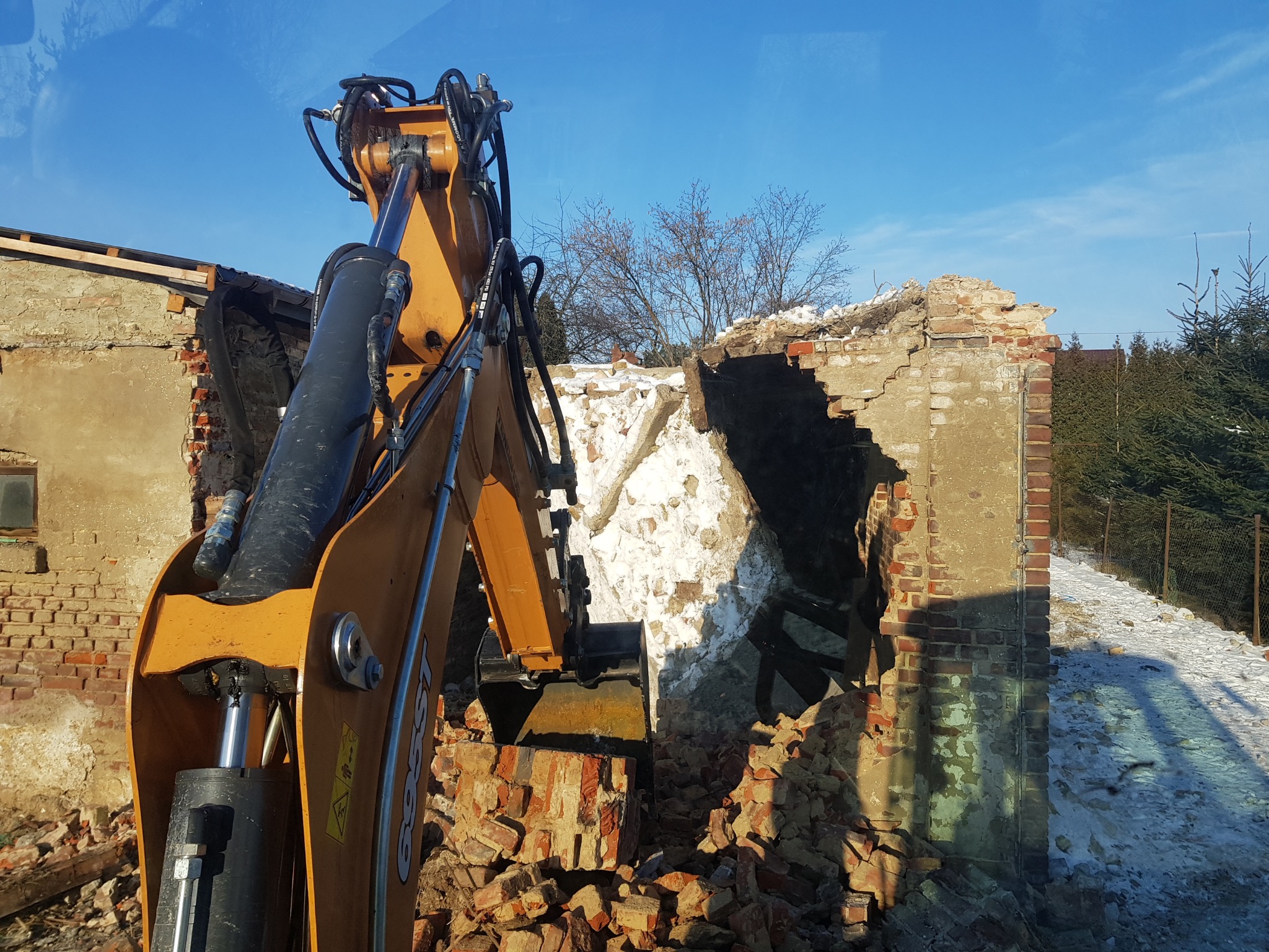 Żółta koparka hydrauliczna kruszy resztki ceglanej ściany budynku w słoneczny dzień, z widocznym śniegiem i drzewami w tle.