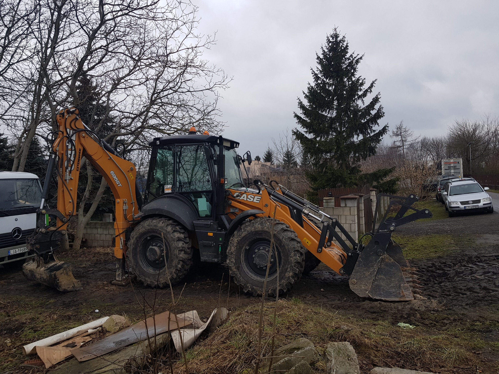 Pomarańczowo-czarna koparko-ładowarka CASE 695ST zaparkowana na błotnistym terenie w pochmurny dzień, widoczny samochód dostawczy Mercedes i osobowy Volkswagen w tle.