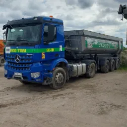 Niebieska ciężarówka z naczepą, z logo firmy EKO-TOM, stojąca na placu z ubitej ziemi, pod pochmurnym niebem. Na naczepie widoczne zielone grafiki i napisy. Na kabinie naklejki informacyjne.