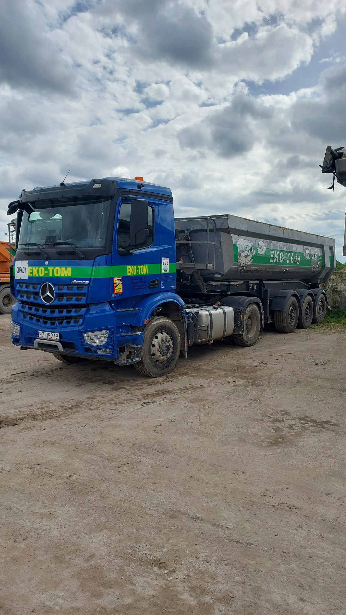 Niebieska ciężarówka z naczepą, z logo firmy EKO-TOM, stojąca na placu z ubitej ziemi, pod pochmurnym niebem. Na naczepie widoczne zielone grafiki i napisy. Na kabinie naklejki informacyjne.