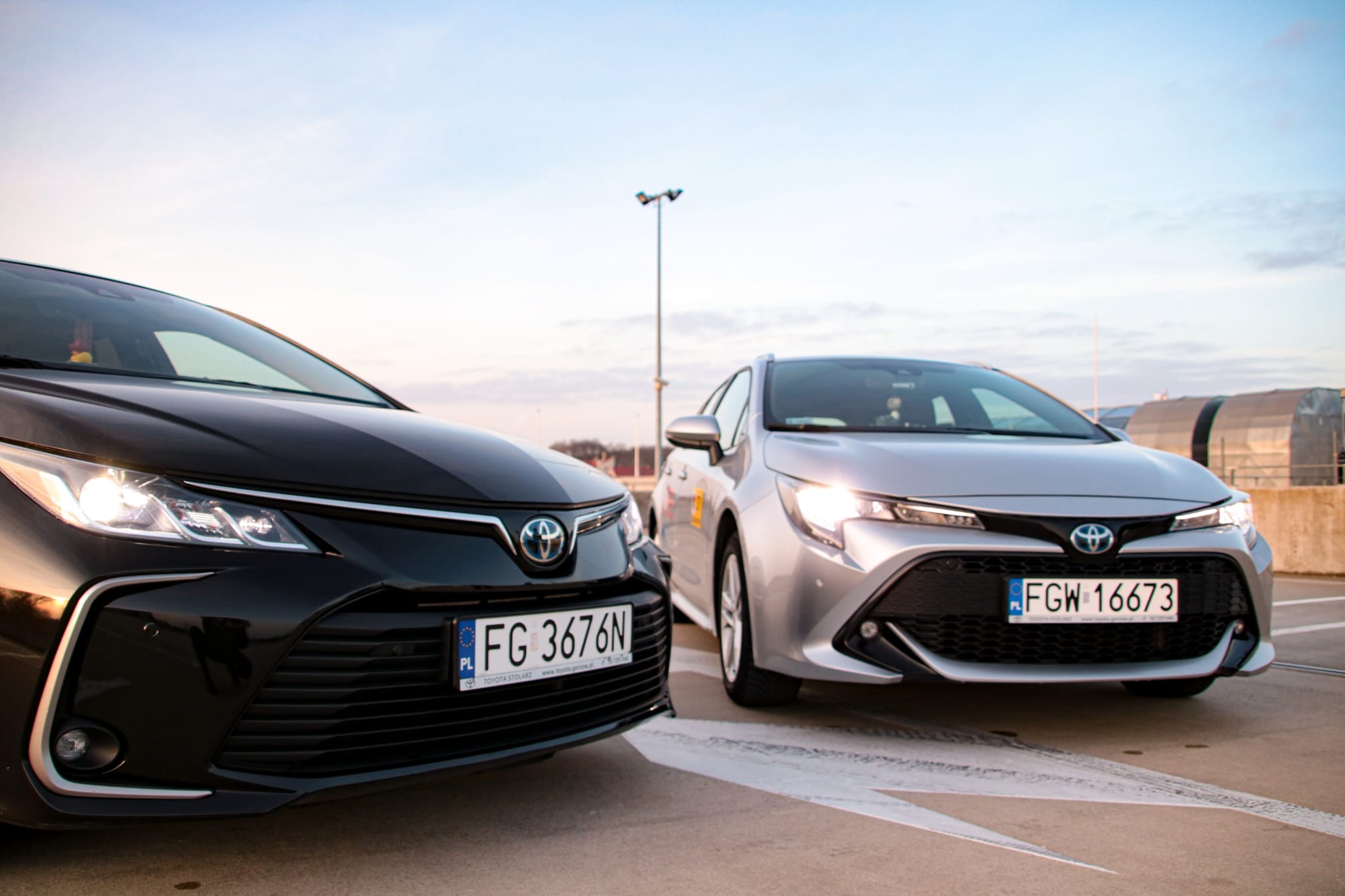 Dwa samochody Toyota Corolla, czarny i srebrny, zaparkowane na parkingu z widocznymi tablicami rejestracyjnymi na tle błękitnego nieba.