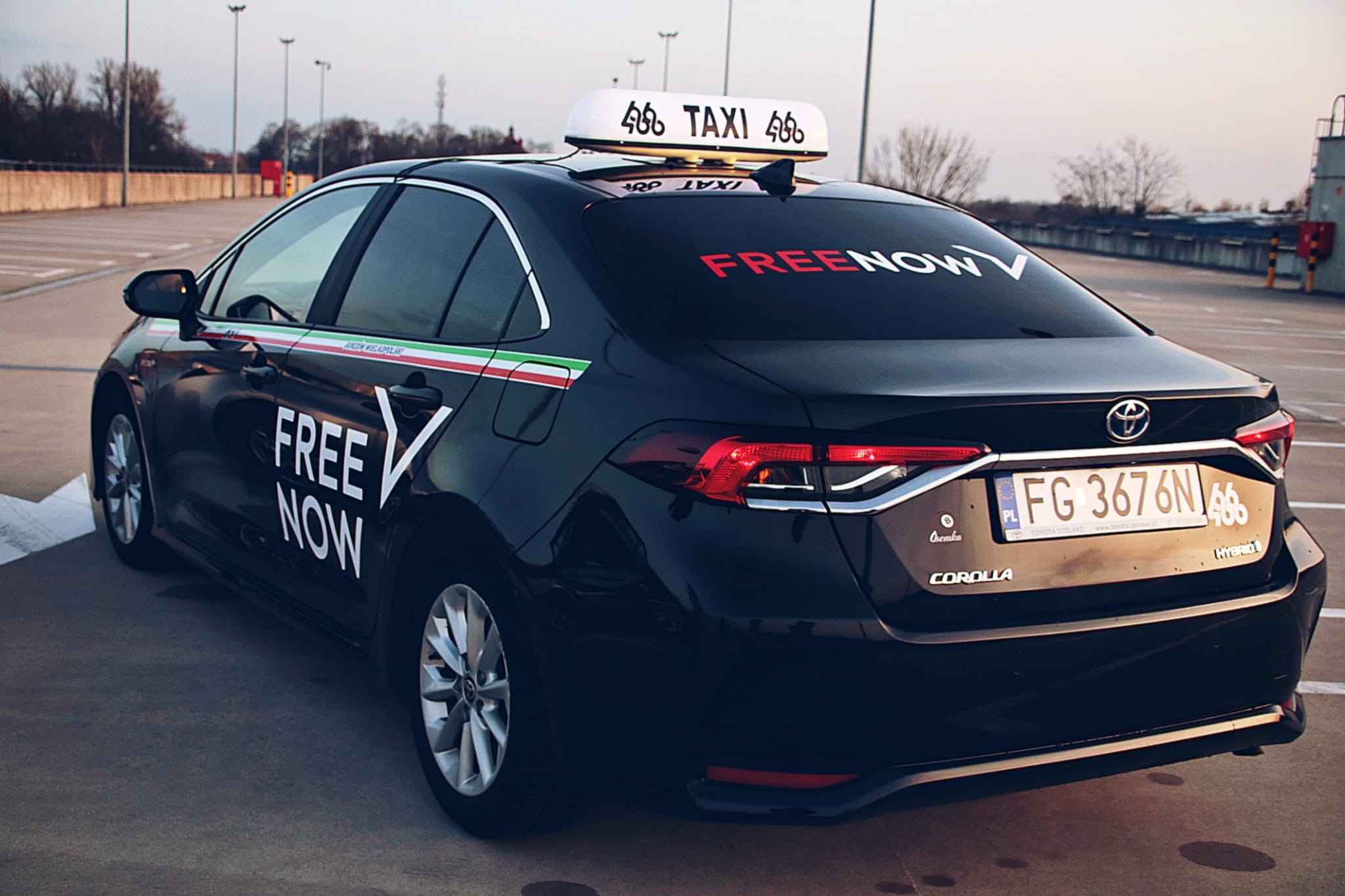Czarny samochód z oznaczeniami firmy przewozowej Free Now i tabliczką Taxi na dachu, zaparkowany na parkingu wielopoziomowym; widoczny numer rejestracyjny, logo Toyoty i oznaczenie Hybrid.