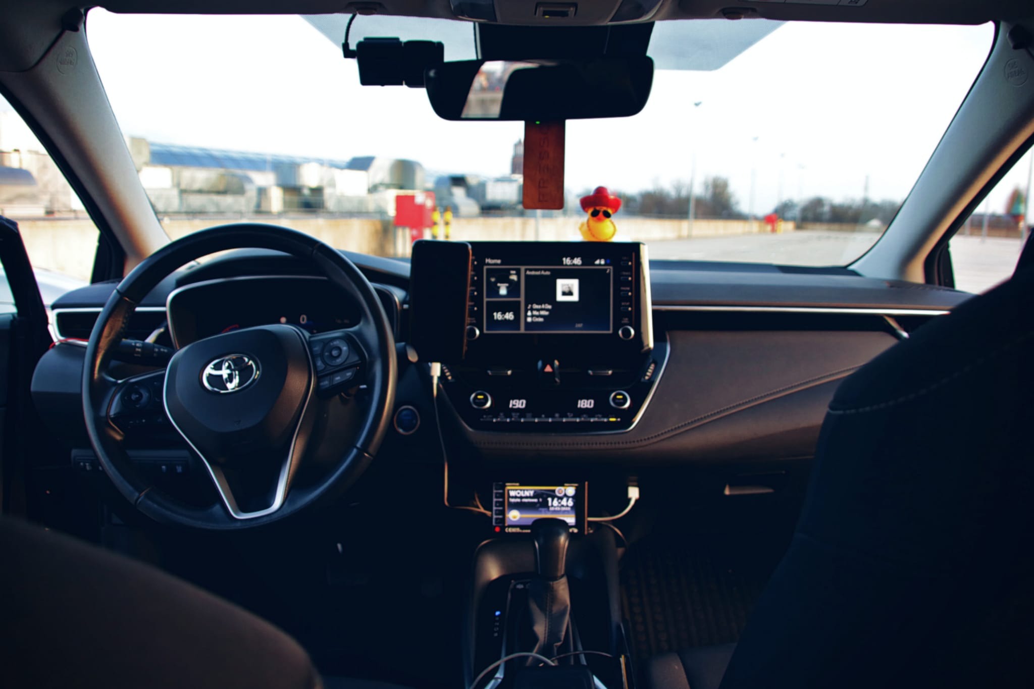 Wnętrze samochodu Toyota Corolla z widocznym kokpitem, kierownicą, ekranem multimedialnym z włączonym Android Auto, kamerą samochodową i gumową kaczką w kapeluszu na desce rozdzielczej.