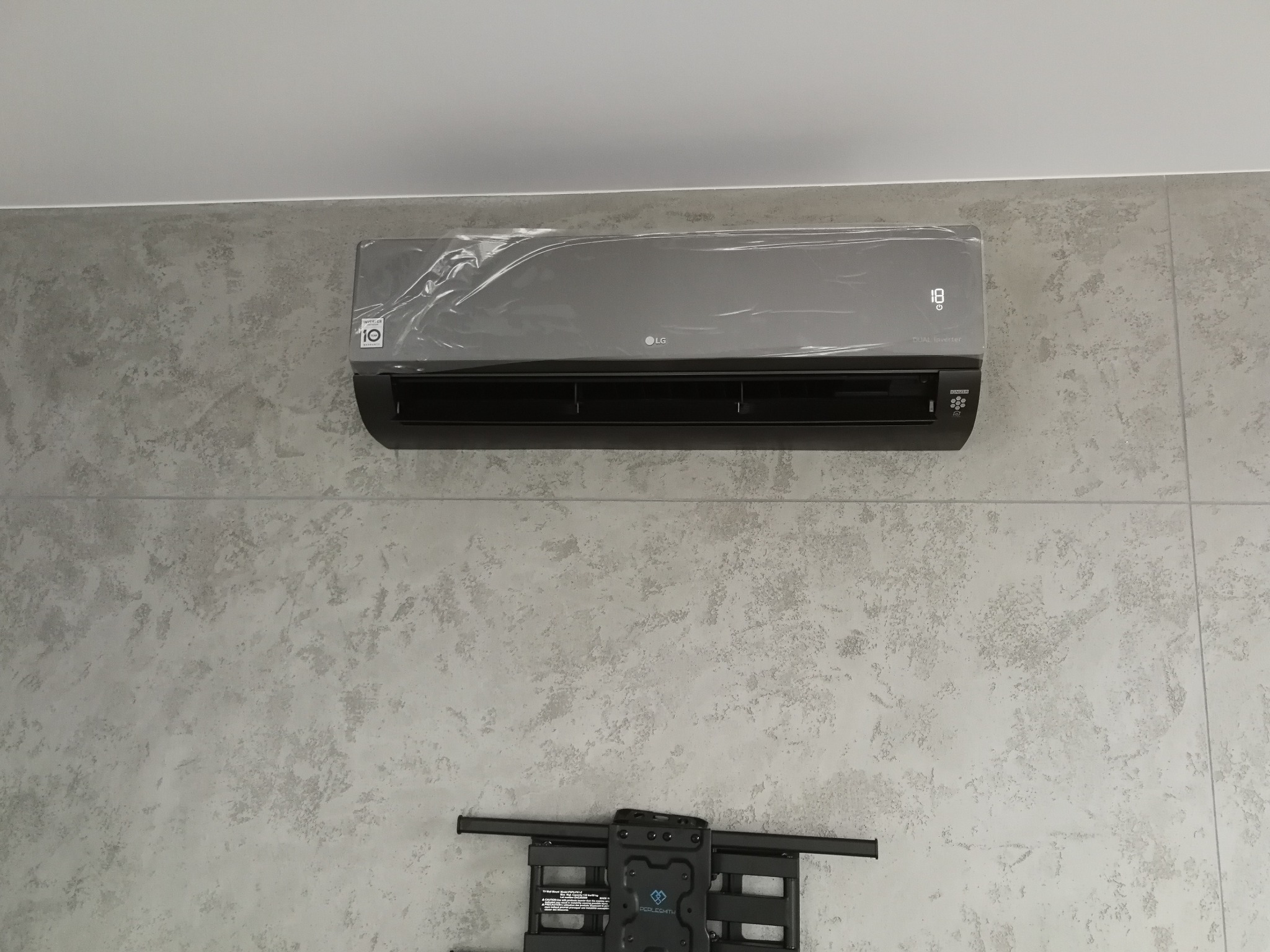 Nowy, szary klimatyzator LG, jeszcze w folii ochronnej, zamontowany na ścianie o teksturze betonu.