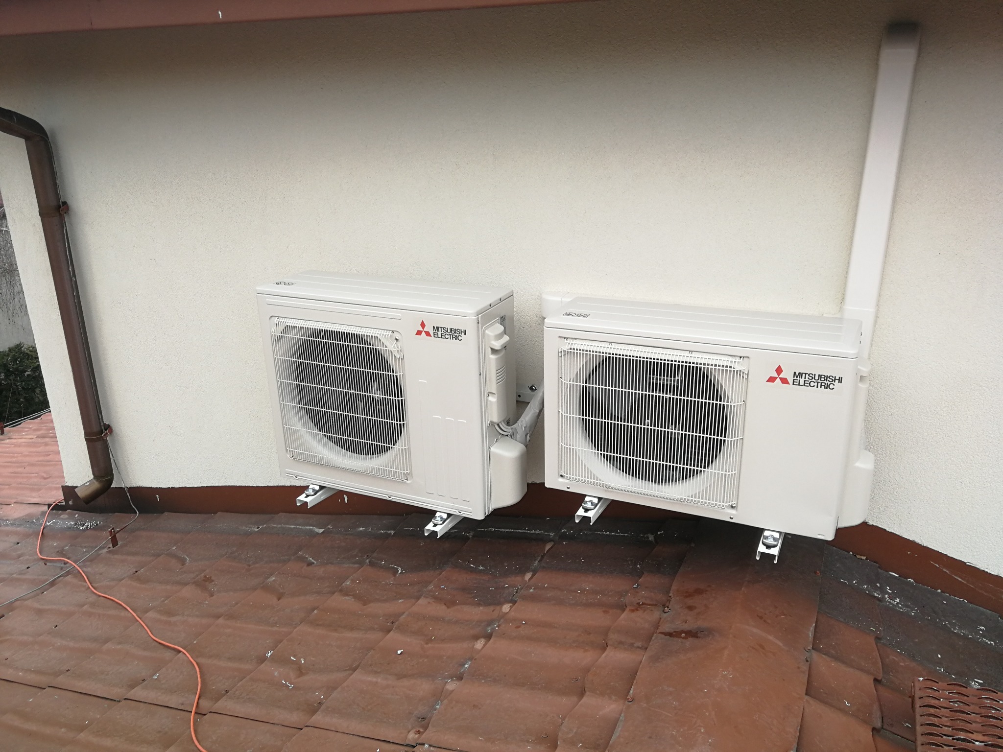 Dwa zewnętrzne moduły klimatyzacji Mitsubishi Electric zamontowane na dachu pokrytym blachą, z widocznymi przewodami i rurami odprowadzającymi skropliny.