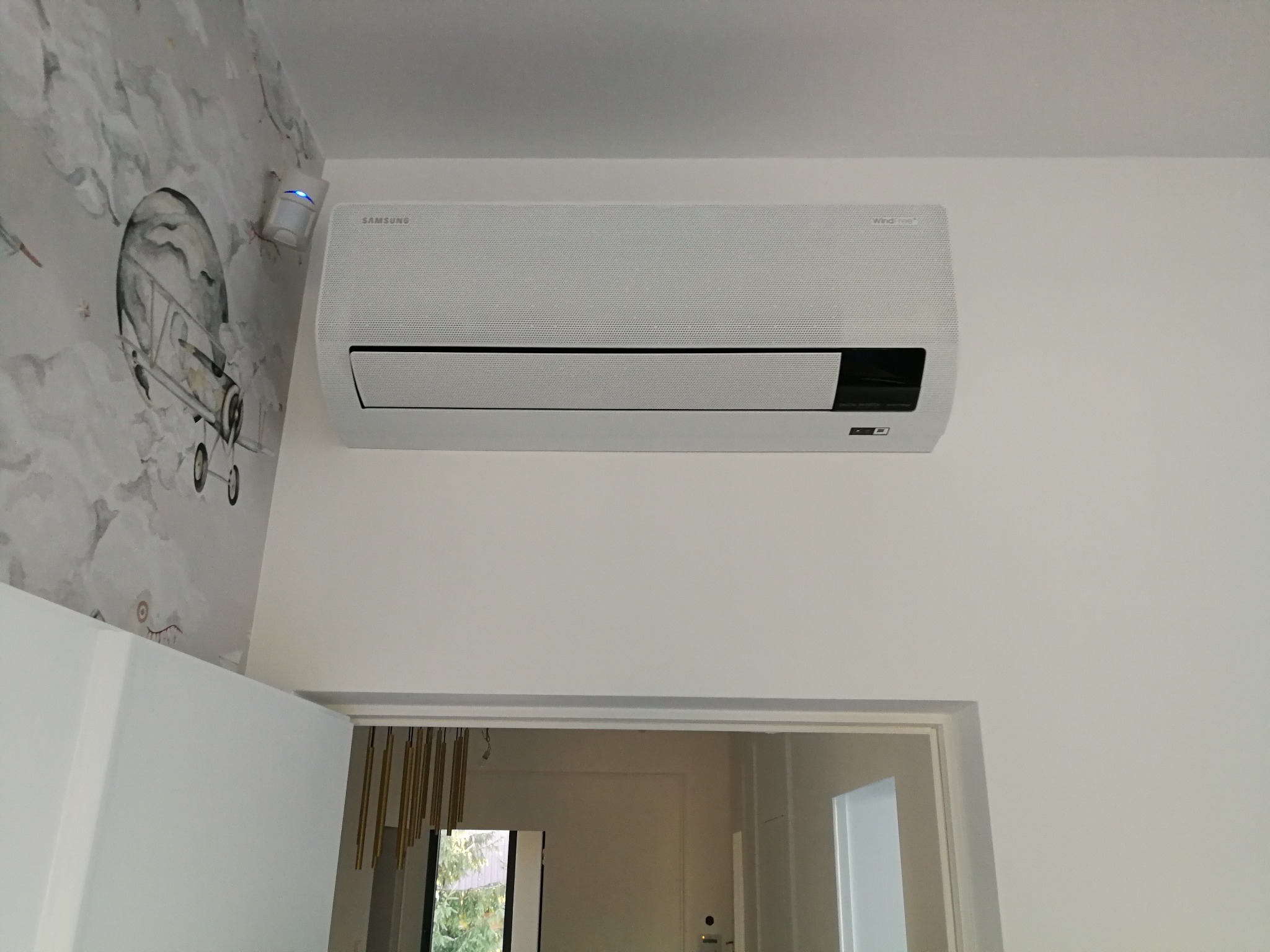 Biały klimatyzator Samsung WindFree zamontowany na ścianie w pokoju dziecięcym z tapetą w balony i samoloty, widoczny czujnik ruchu.