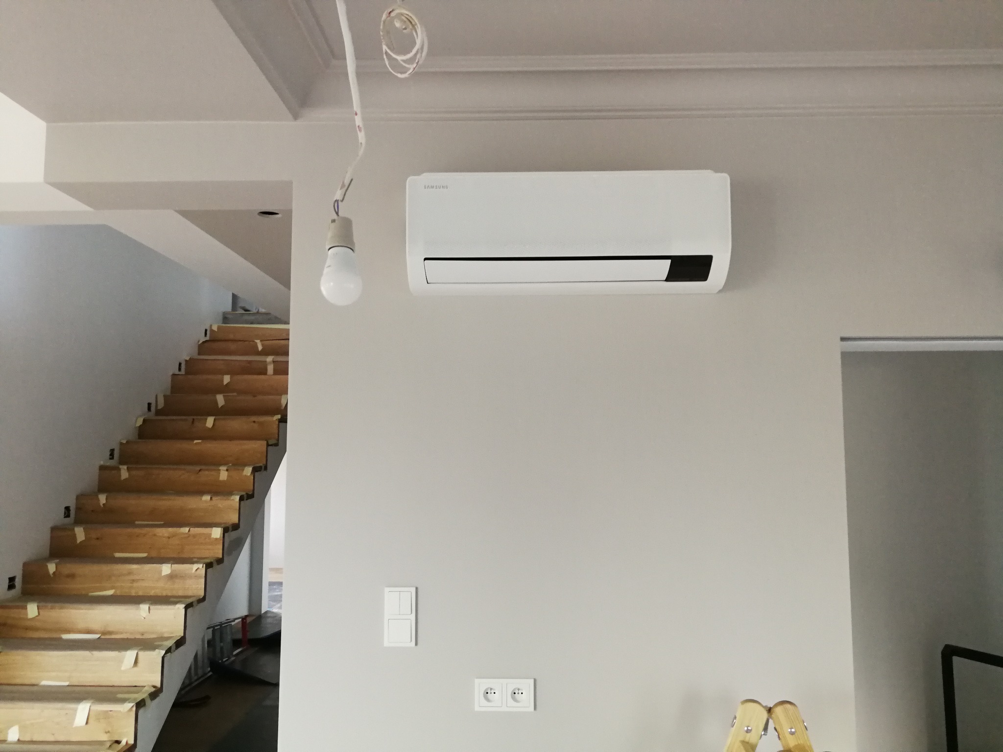 Biały klimatyzator Samsung zamontowany na szarej ścianie w nowym domu, widoczne schody w surowym stanie, żarówka na kablu i gniazdka elektryczne.