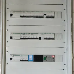 Rozdzielnica elektryczna z zamontowanymi zabezpieczeniami, wyłącznikami i modułem pomiarowym z wyświetlaczem, widoczne przewody w dolnej części szafki, ściana w surowym stanie.