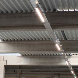 G-install - Widok z dołu na metalową korytkę kablową z zamontowanymi lampami LED, zawieszoną pod betonowym stropem w hali przemysłowej.