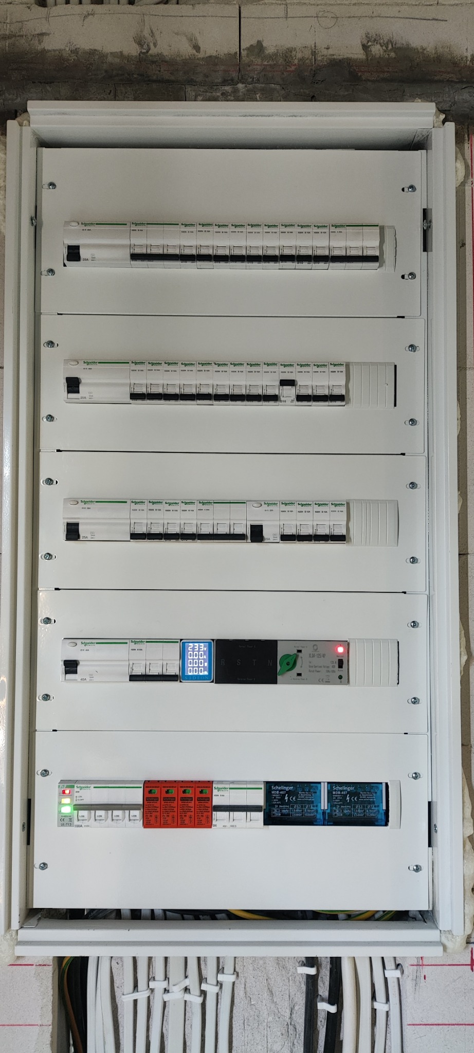 Rozdzielnica elektryczna z zamontowanymi zabezpieczeniami, wyłącznikami i modułem pomiarowym z wyświetlaczem, widoczne przewody w dolnej części szafki, ściana w surowym stanie.