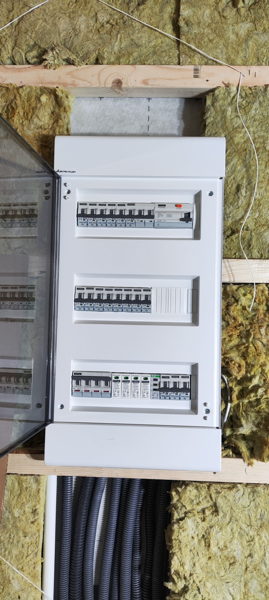 Otwarta biała skrzynka rozdzielcza z bezpiecznikami marki Schillinger zamontowana w ścianie z izolacją z wełny mineralnej, widoczne szare rury karbowane z kablami.