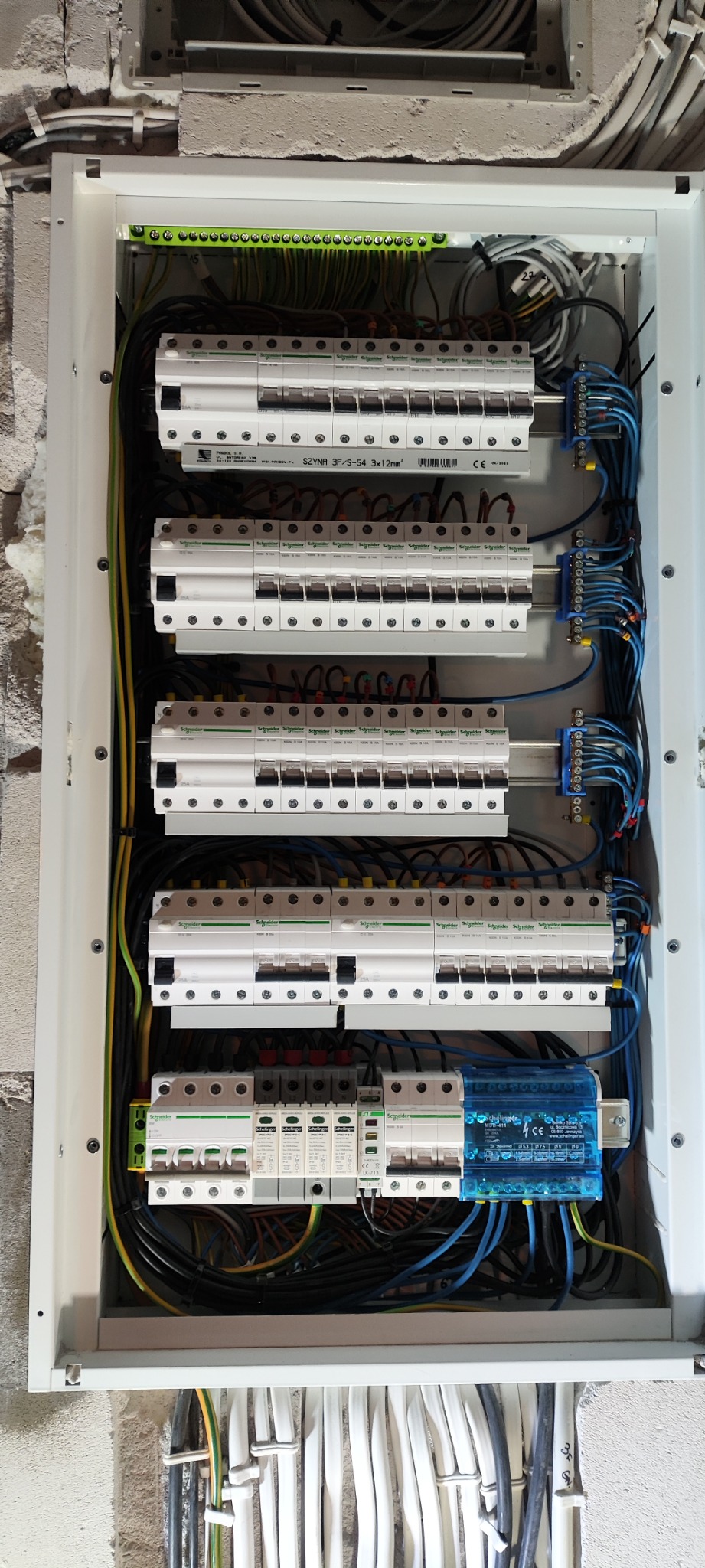 Rozdzielnia elektryczna z zamontowanymi bezpiecznikami, warystorami i listwami zaciskowymi w białej obudowie, widoczne przewody w peszlach wystające ze ściany.