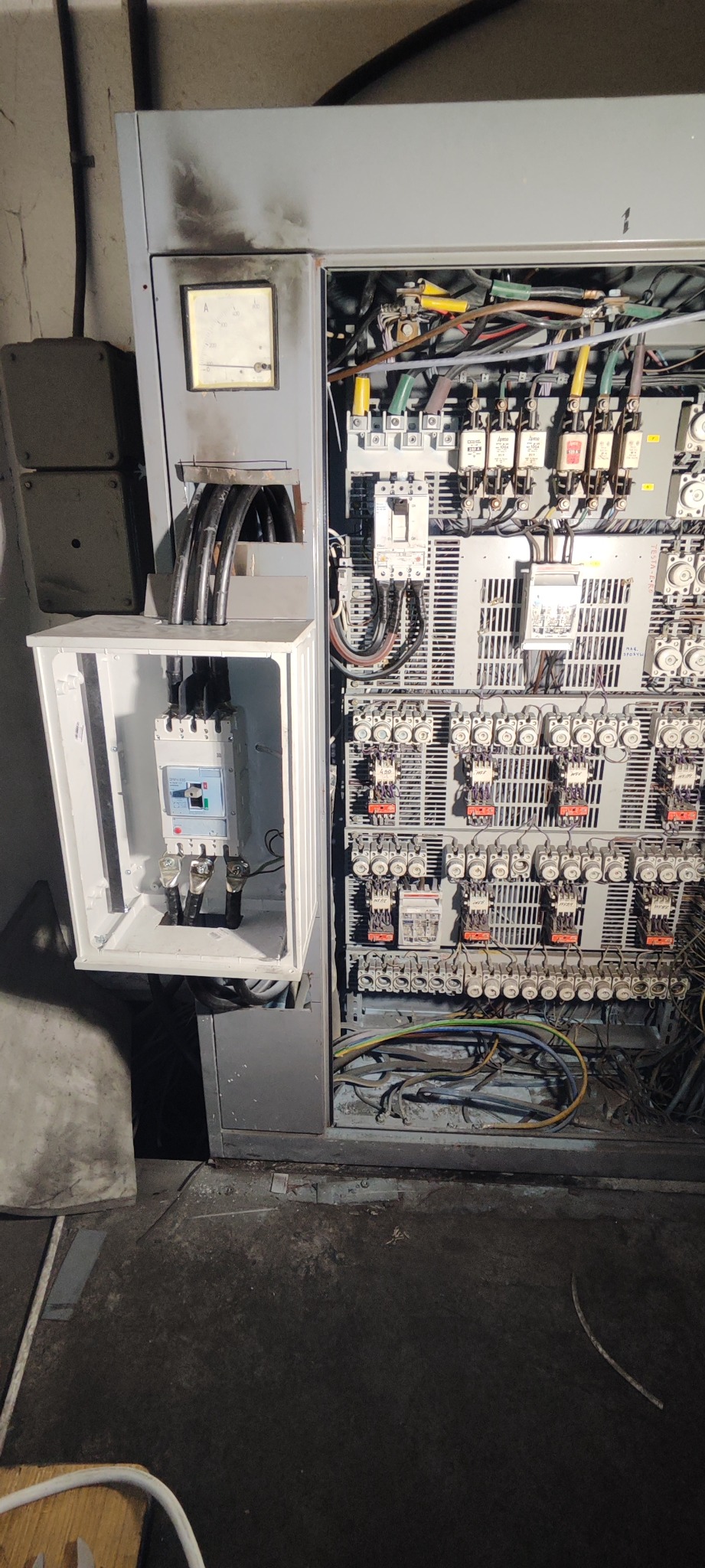 Otwarta szafa elektryczna z nowym wyłącznikiem w obudowie, widoczne liczne kable, bezpieczniki i elementy sterujące, ślady okopcenia na obudowie szafy.