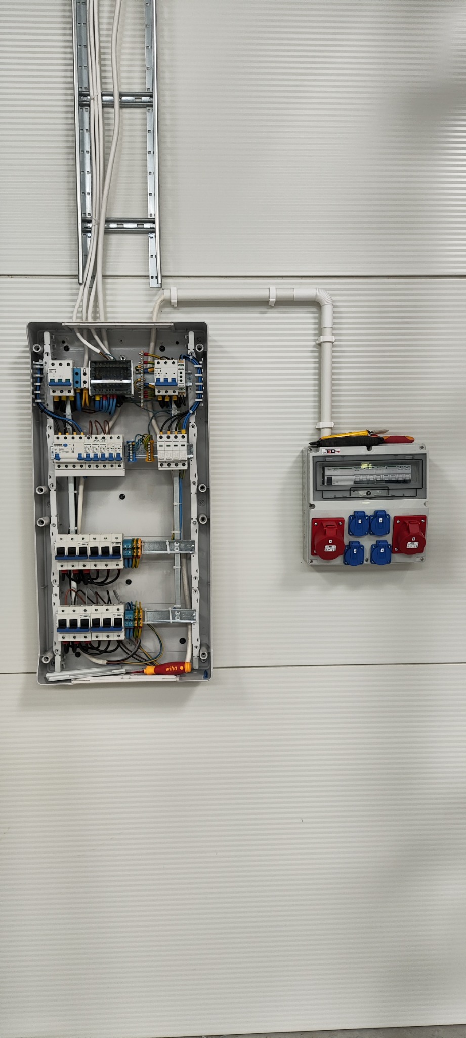 Otwarta skrzynka rozdzielcza z widocznymi bezpiecznikami i przewodami, obok zamknięta skrzynka z gniazdami siłowymi na ścianie z poziomymi panelami. W dolnej części otwartej skrzynki leży śrubokręt...