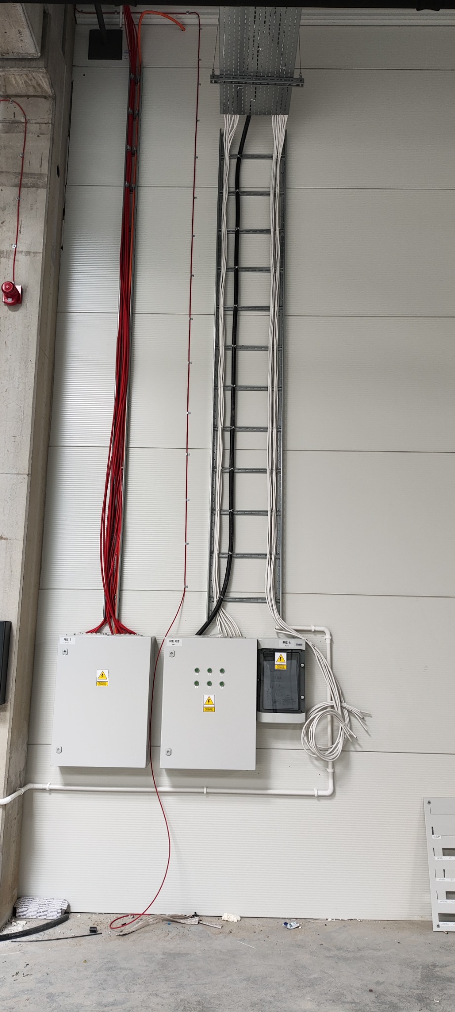 Rozdzielnice elektryczne natynkowe z przewodami prowadzonymi w korytach kablowych i po ścianie w hali przemysłowej.