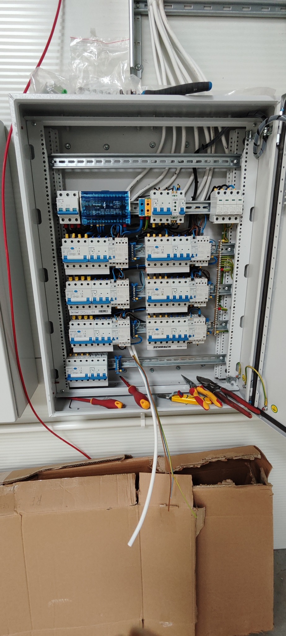 Otwarta szafa elektryczna z zamontowanymi bezpiecznikami, zabezpieczeniami i przewodami. Na dole szafy leżą dwa śrubokręty i kombinerki. Dolna część zdjęcia zasłonięta kartonem.