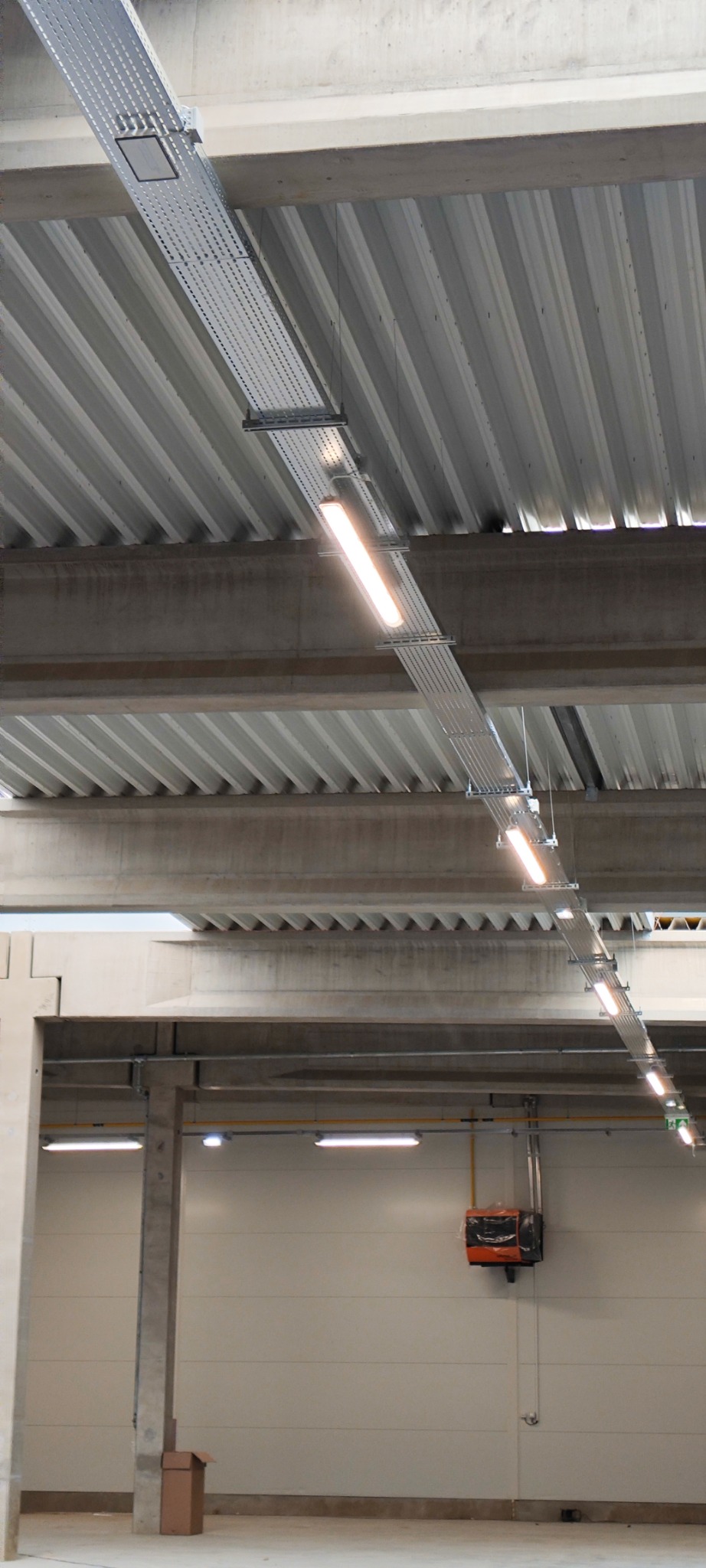 Widok z dołu na metalową korytkę kablową z zamontowanymi lampami LED, zawieszoną pod betonowym stropem w hali przemysłowej.