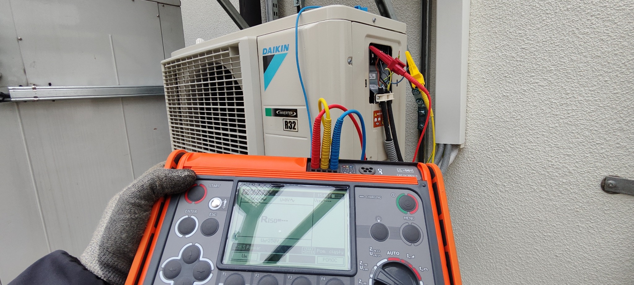 Pomiar parametrów elektrycznych klimatyzatora Daikin R32 Inverter za pomocą pomarańczowego miernika z kolorowymi przewodami pomiarowymi.