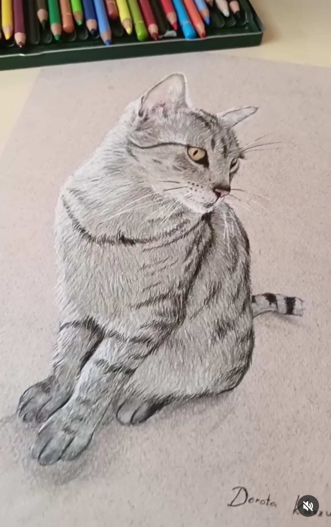 Ręcznie rysowany portret siedzącego, pręgowanego kota w odcieniach szarości, wykonany kredkami na beżowym papierze, z widocznym pudełkiem kredek w tle.