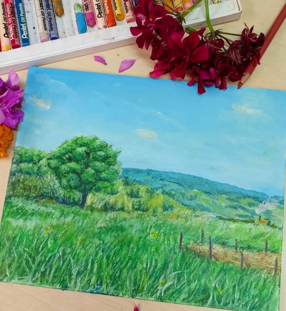 Pejzaż namalowany pastelami olejnymi przedstawiający zielone pole, drzewa i wzgórza pod błękitnym niebem, z pudełkiem pasteli i kwiatami w tle.