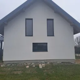 Świeżo otynkowana elewacja domu jednorodzinnego z trzema oknami o czarnych ramach; na trawniku leżą bloczki betonowe. Widok z przodu, pochmurne niebo.