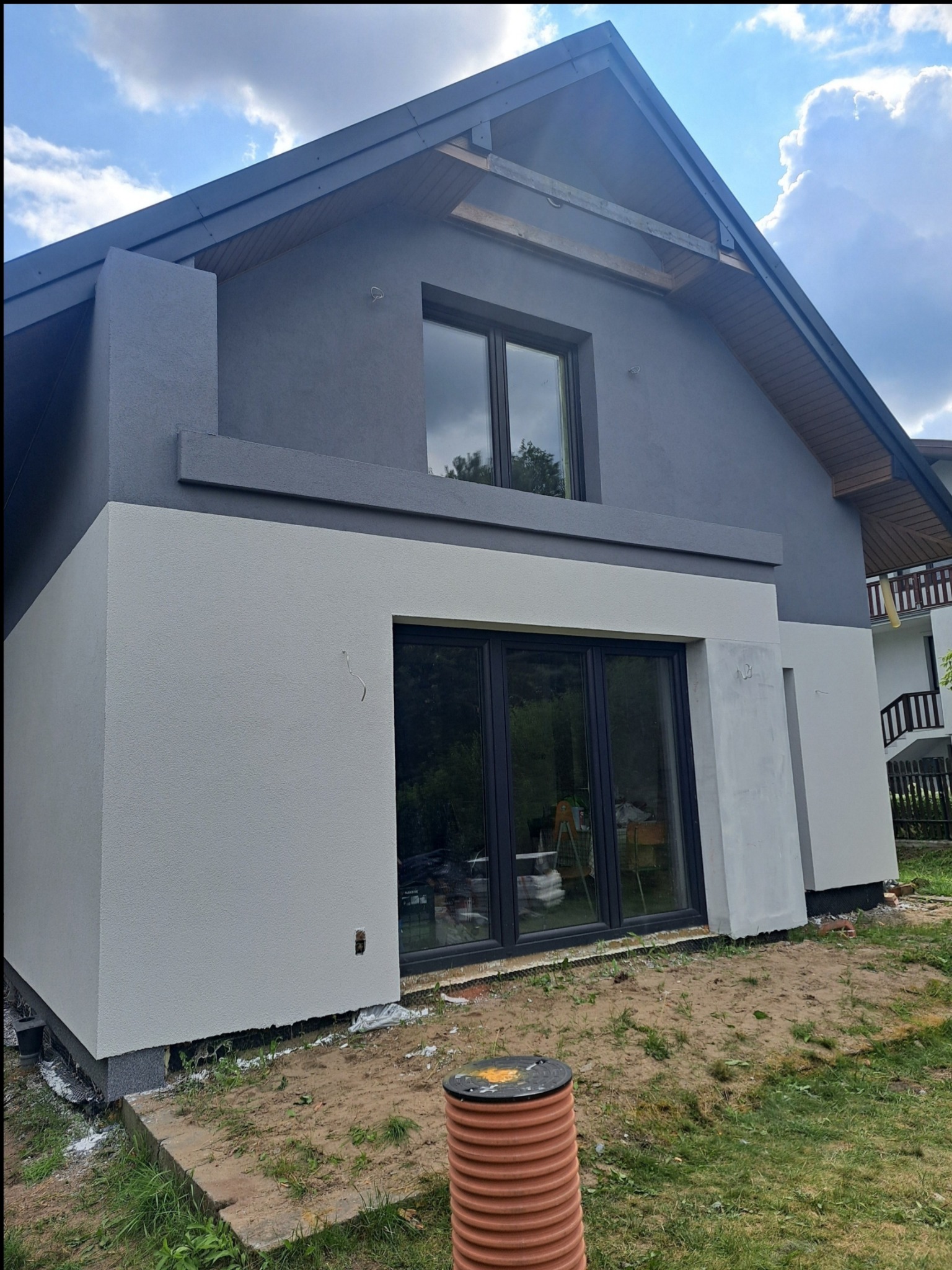 Dwukolorowa elewacja domu jednorodzinnego w odcieniach szarości i bieli. Widoczne okna z ciemnymi ramami i fragment ogrodu. Częściowo otynkowana ściana.