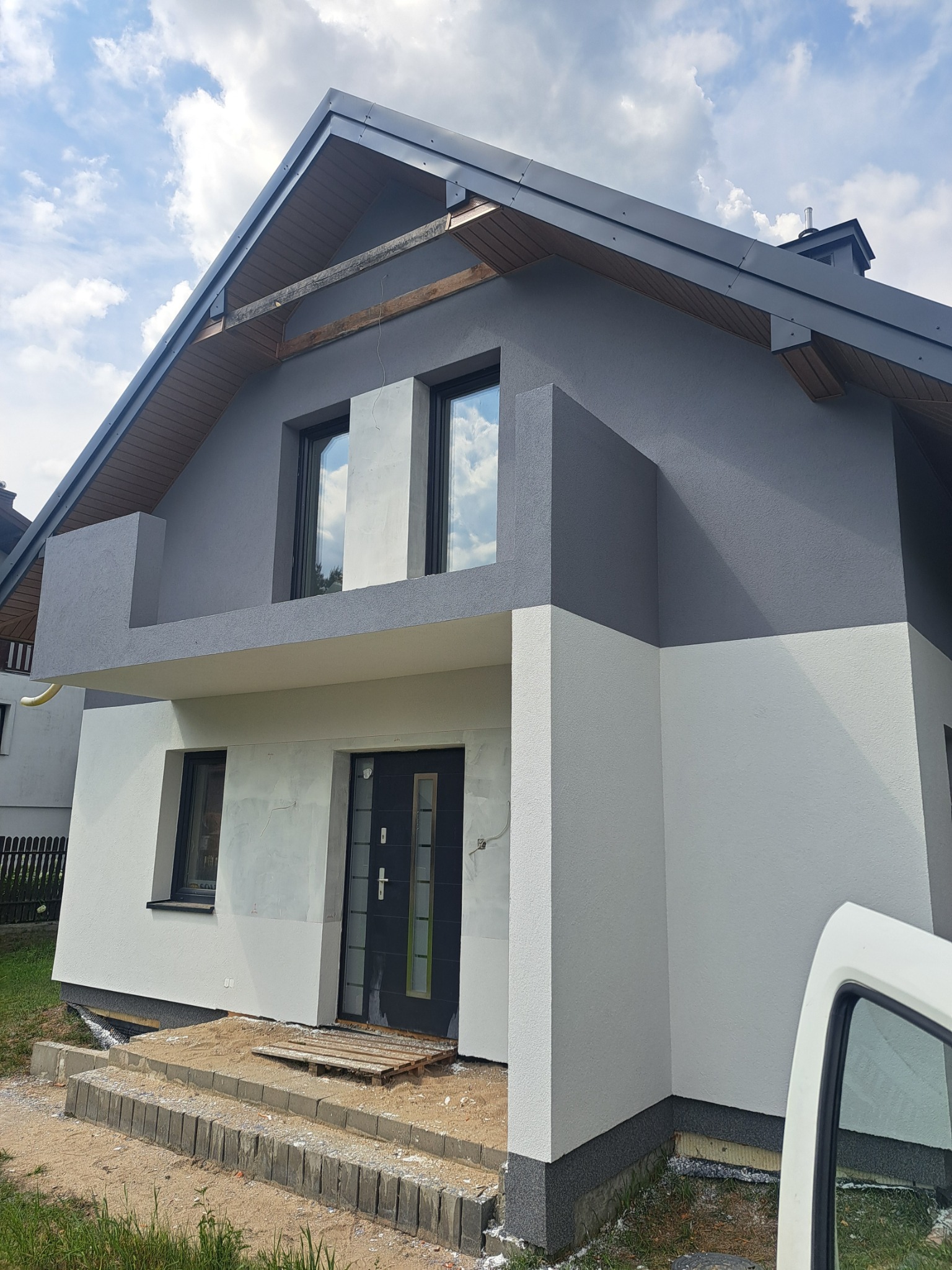 Dwukolorowa elewacja domu jednorodzinnego w odcieniach szarości i bieli, z widocznymi elementami wykończeniowymi i schodami wejściowymi. Częściowo widoczny samochód.