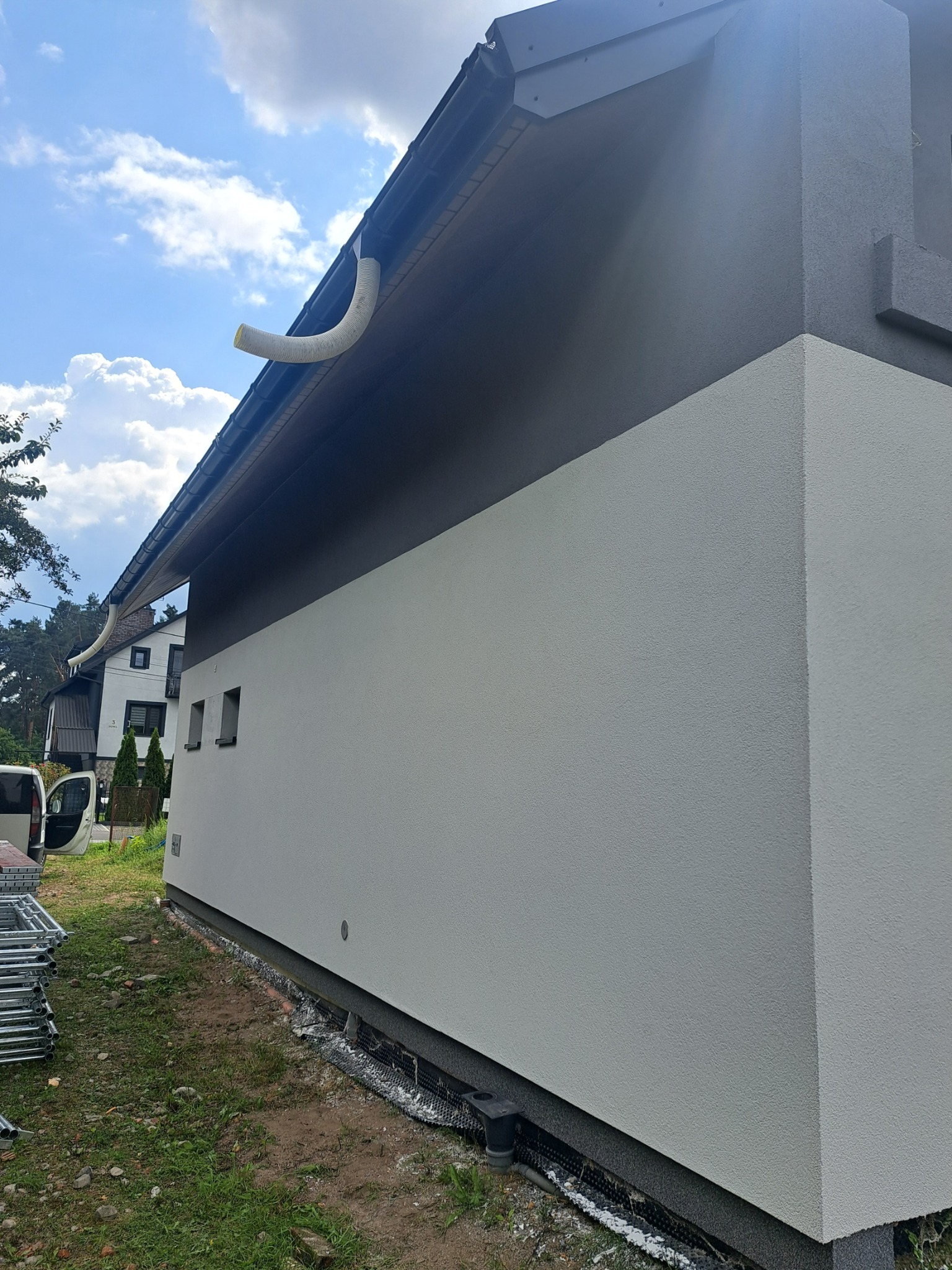 Dwukolorowa elewacja domu jednorodzinnego w słoneczny dzień. Widoczne rynny, fragment rusztowania i trawnik. Jasna, gładka dolna część i ciemnoszara górna.