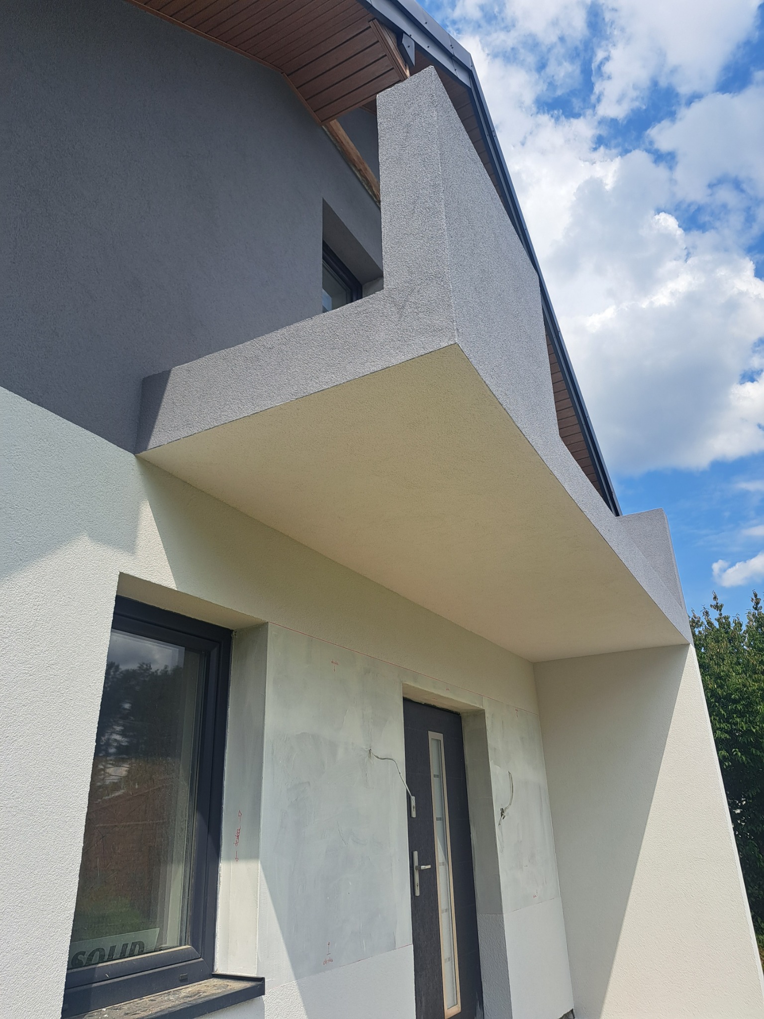 Szara elewacja domu z widocznym oknem i drzwiami wejściowymi, fragment ściany w trakcie prac wykończeniowych, błękitne niebo w tle. Ujęcie z dołu.